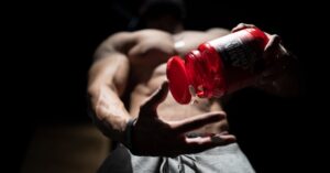 Shirtless muscular man pouring protein supplements from a bottle, showcasing fitness and strength.
